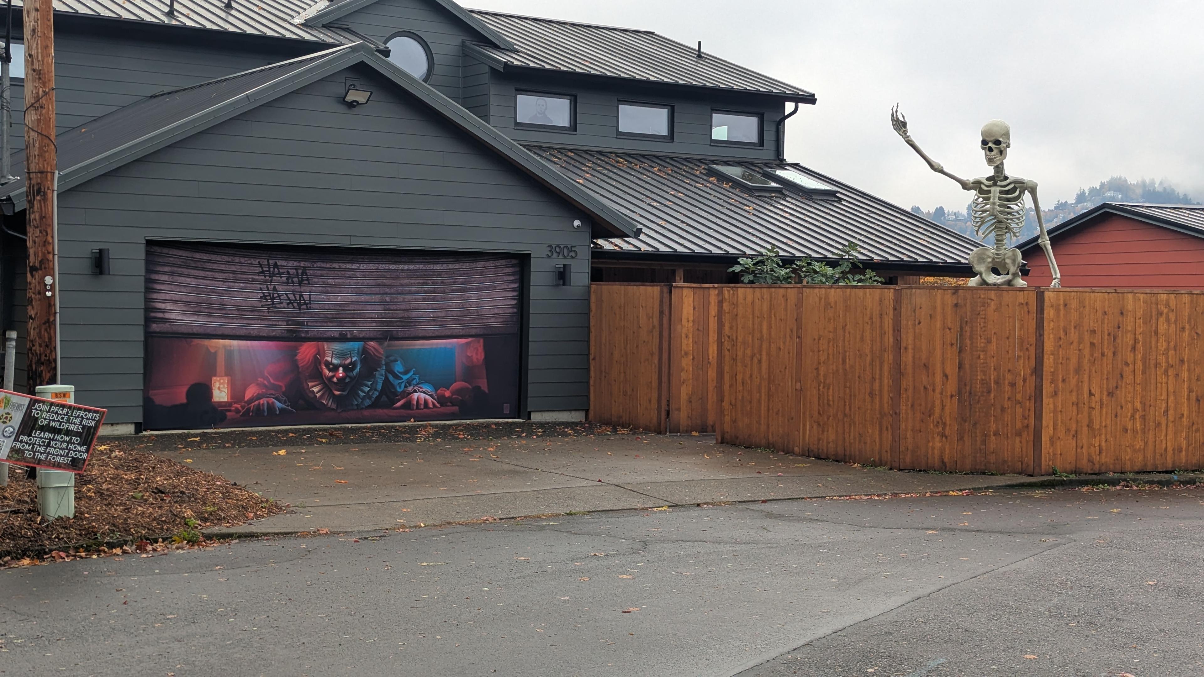 A wide view of a house to the left. The garage is painted with a mural of the clown from IT. To the right is a 12-ft skeleton standing in the yard. One arm raised as if in dramatic speech. The backdrop is a foggy Pacific Northwest day.