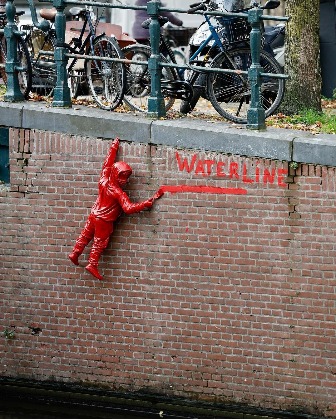 Red sculpture installation by street artist James Colomina in Amsterdam, Netherlands, depicting a child in a hooded raincoat clinging to a canal wall while spray-painting the word “WATERLINE” in red. The artwork, placed just above the water level beneath parked bicycles, comments on rising sea levels and climate change in a city known for its canals.