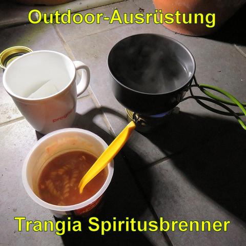 Ein Trangia Spiritusbrenner steht auf einem gefliesten Boden. Darauf ein kleiner schwarzer Topf, in dem Wasser kocht, mit Dampf, der aufsteigt. Daneben eine weiße Tasse sowie ein geöffneter Becher mit roter Suppe und einer gelben Plastiklöffel darin. Der Spiritusbrenner hat grüne Klappgriffe am Topf, während die Tasse leer zu sein scheint. Die Szene wirkt wie ein improvisiertes Outdoor-Kochsetup.