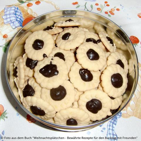 Auf dem Tisch steht eine Blechdose mit hellgelben Marzipan-Orangen-Blumen. In der Mitte von jedem Keks ist ein Klecks Zartbitterkuchenglasur.