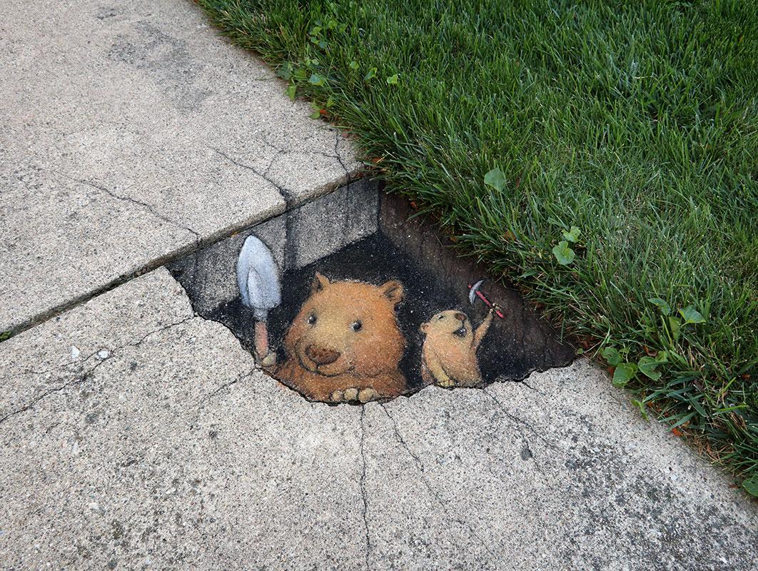 Chalk art by David Zinn on a cracked sidewalk depicts two small burrowing animals peeking out from a hole in the pavement. One holds a tiny shovel and the other raises a miniature pickaxe, as if they’re excavating beneath the concrete. The surrounding grass and worn sidewalk frame the illusion, blending street and imagination in Zinn’s signature playful style.
