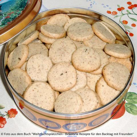 Auf dem Tisch steht eine Blechdose mit vielen kleine gelbbräunlichen Marzipan-Mandel-Talern. Die Kekse haben von den gemahlenen Mandeln eine gesprenkeltes Aussehen.