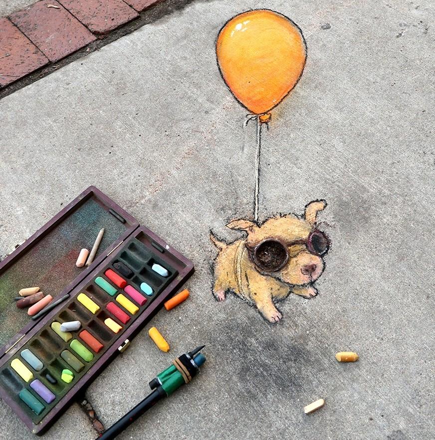 A whimsical sidewalk chalk drawing by artist David Zinn showing a tiny yellow puppy wearing oversized, round, metal-look goggles, illustrated as if itโs floating upward while holding onto a bright orange balloon tied to its back. The artwork is drawn directly on a concrete pavement, with scattered pastel chalk sticks and an open chalk box beside it, emphasizing Zinnโs playful, imaginative style. The piece is titled โPup in Flightโ and was created on a street corner where the chalk drawing integrates naturally with the texture of the sidewalk.
