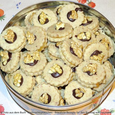 Viele kleine gefüllte Mürbeteigplätzchen mit aufgeklebtem Walnussstück liegen in einer Blechdose auf dem Tisch.