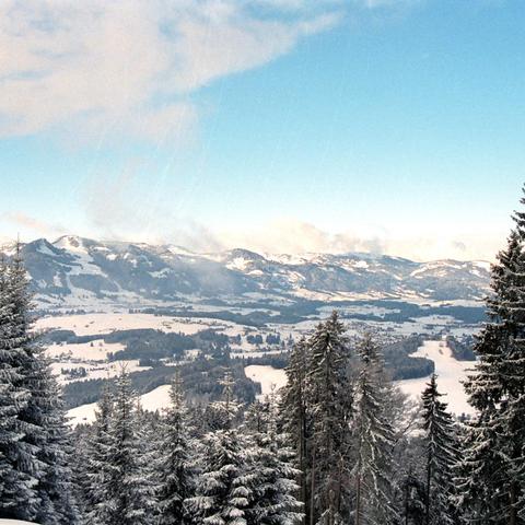 Dieses Bild zeigt eine schneebedeckte Landschaft in den Alpen. Im Vordergrund stehen hohe Nadelbäume, deren Äste schwer von Schnee beladen sind. Dahinter öffnet sich die Landschaft zu einer weiten Ebene, die von Schneeflächen und dunklen Baumgruppen durchbrochen wird. Mehrere Hügel erheben sich in der Mitte des Bildes und am Horizont zeichnen sich die Umrisse von höheren Bergen ab. Die Wolken am blauen Himmel sind spärlich und lassen viel Sonnenlicht durch, welches die Schneeflächen zum Glänzen…