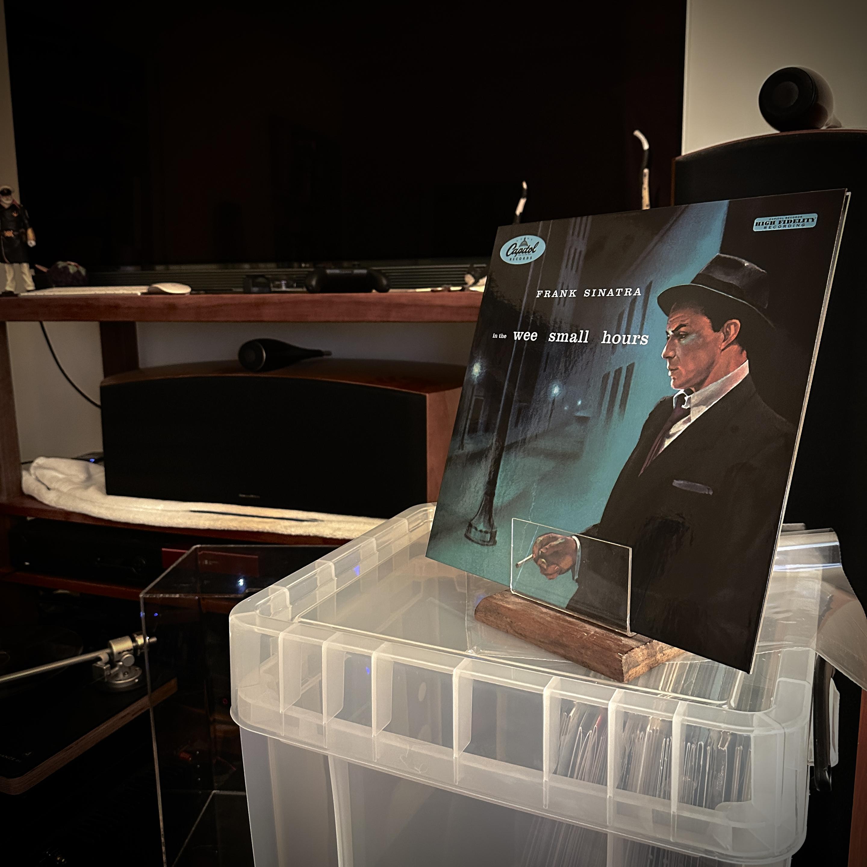 The current mess of my Now Playing setup (I swear I’ll clean this up before the end of the year). Atop a translucent white LP crate box is my LP prop and in it is Frank Sinatra’s ‘In the Wee Small Hours’ LP. It shows a painting(?) of a young Frank leaning against the corner of a building, facing toward the title, smoking a cigarette. Over his shoulder is a blue quiet street scene. A lamp post is visible in the midground, and additional street lamps can be seen in the distance. The painting has an abstract dreamy quality about it which gives the viewer the notion of longing, and sadness. Frank’ expression is contemplative.