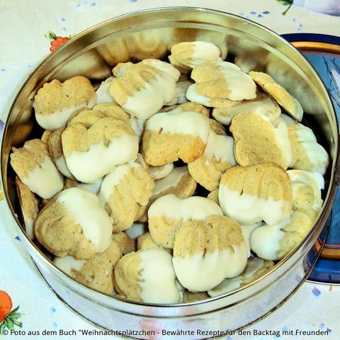 Auf dem Tisch steht eine offene Blechdose mit goldgelbem Weihnachtsspritzgebäck in S-Form. An einer Seite haben die Plätzchen eine weiße Schokoladenkante.