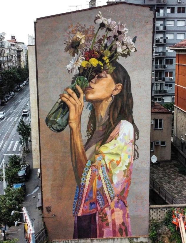 A large mural titled “Thirst for Nature” by Artez painted on the side of a tall residential building in Belgrade, Serbia. The artwork shows a woman with long dark hair holding a green glass bottle filled with wildflowers, lifting it to her face as if drinking in their scent. She wears a colorful patterned garment with soft pastel tones. The mural covers the entire facade and stands out against the urban surroundings of apartment buildings and a busy street lined with trees.