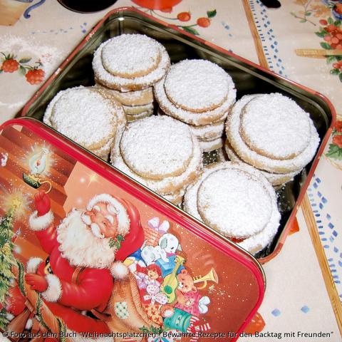 Auf dem Tisch steht eine eckige Blechdose mit Weihnachtsmannaufdruck auf dem Deckel. Der Deckel ist geöffnet und man sieht sechs mit Puderzucker bestreute gestapelte Mürbeteigplätzchen-Säulen.