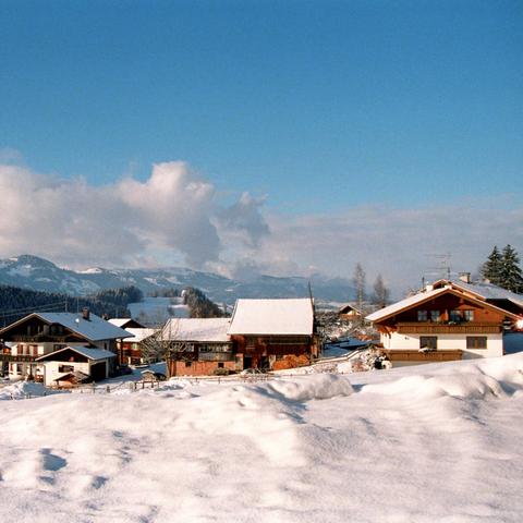 Das Bild zeigt eine Nahaufnahme eines Dorfes im Winter in den Alpen. Mehrere Chalets und traditionelle Gebäude mit schneebedeckten Dächern sind sichtbar. Die Architektur ist typisch alpin mit massiven Holzkonstruktionen und großen Dächern, die den Schneefall aushalten können. Im Hintergrund sind verschneite Berge und ein blauer Himmel mit wenigen Wolken zu sehen, was auf klares Wetter hindeutet. Die Umgebung ist friedlich und die Schneedecke reflektiert das Sonnenlicht, was die Szene besonders …