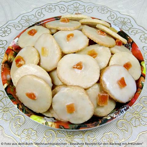 Auf einem Deckchen mit Goldkante steht ein Pappteller mit Weihnachtsaufdruck. In dem Teller liegt ein Stapel helle Orangen-Plätzchen mit Zuckerguss und in der Mitte ein Stück kandierte Orange.