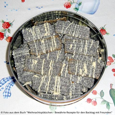 Auf dem Tisch steht eine Blechdose mit rechteckigen gräulichen Mohn-Orangen-Plätzchen, die quer gestreift sind mit Vanillekuchenglasur.