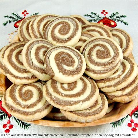 Auf einer Weihnachtstischdecke aus Stoff mit aufgestickten roten Kerzen und Tannengrün steht ein holzfarbener eckiger Teller mit Zwei-Farben-Schnecken. Das Spiralmuster ist hellgelb-hellbraun.