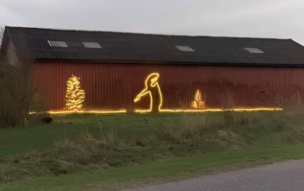 A rural red barn decorated with a glowing yellow light installation showing La Linea silhouette pointing between two illuminated Christmas trees, with a continuous line of lights running along the ground. The humorous outdoor light artwork contrasts festive holiday symbols with crude humor, set against a grassy roadside landscape at dusk. Artist unknown, location unknown.