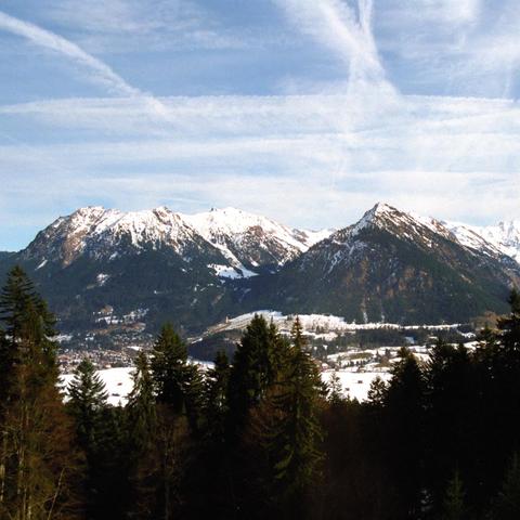 Blick Richtung Oberstdorf vom Berghang. Im Vordergrund stehen dunkelgrüne Tannen, die das schneebedeckte Tal mit verstreut liegenden Häusern einrahmen. Dahinter ragen markante Berggipfel empor, teils schneebedeckt, teils mit dunklen Felsflächen und Wäldern. Der Himmel ist klar und blau, durchzogen von weißen Kondensstreifen, die die Weite der Landschaft unterstreichen.