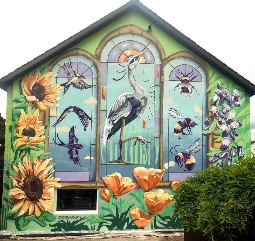 Gable-end mural by Shona Hardie in Edinburgh, Scotland, UK, painted like a three-panel stained-glass window. In the center arch, a tall heron stands in shallow water with reeds behind it, framed by curved window panes. Around it, small birds fly across a blue sky, while oversized bees and insects hover in the side panels. Large sunflowers climb the left edge of the wall, orange poppies bloom along the bottom, and purple foxglove flowers rise on the right, all set against a soft green background on a residential building.