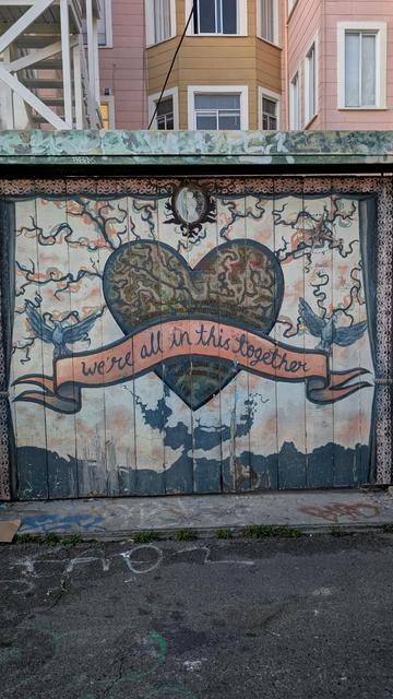 Square painted mural with a heart in the center. A banner in front of the heart reads "We Are All In This Together"