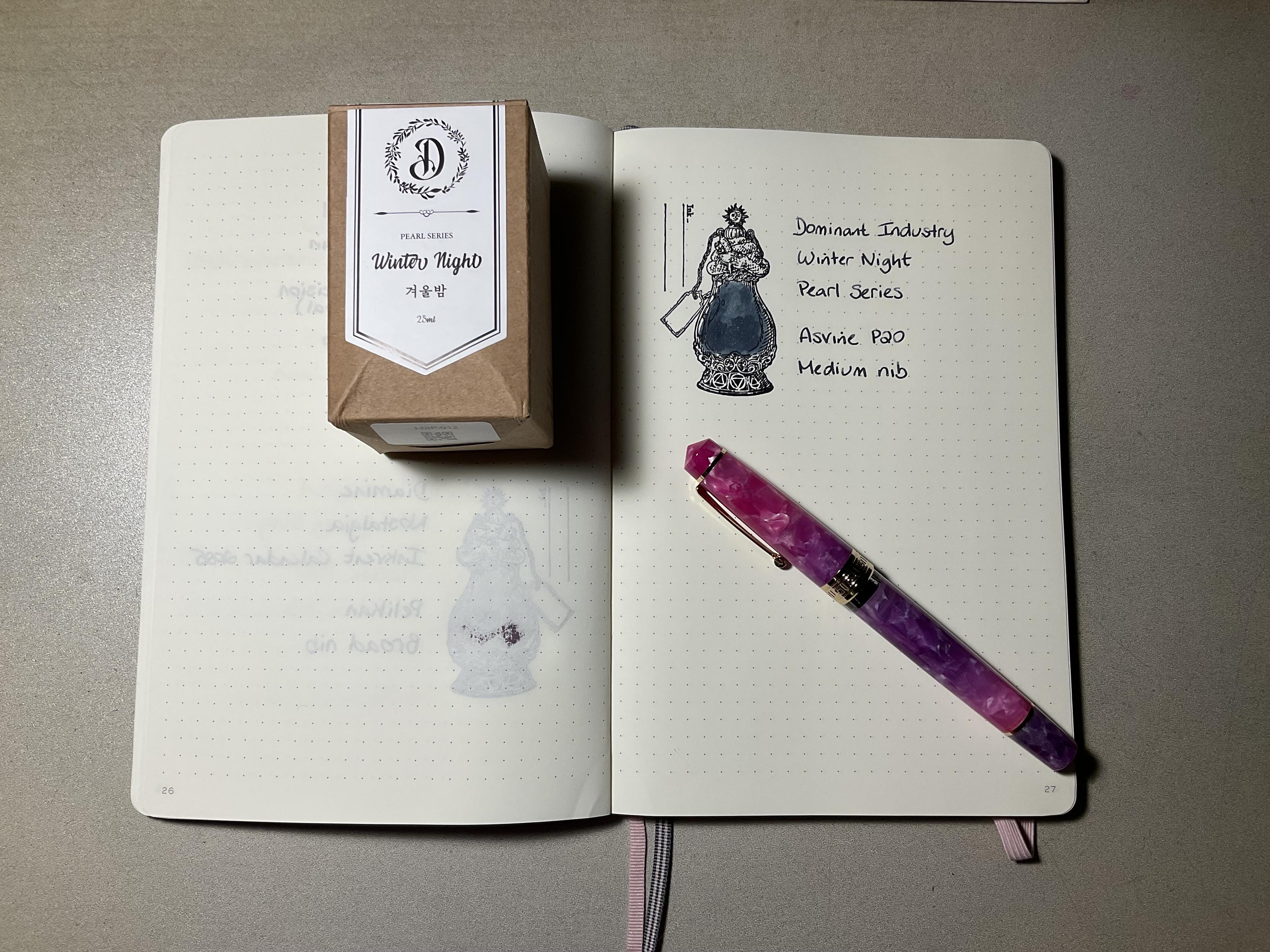 Ink swatch in an open notebook with an ornamental bottle stamp. The colour is dark grey with pearl glitter. The box is on the left side and a pink fountain pen is placed beneath the swatch.