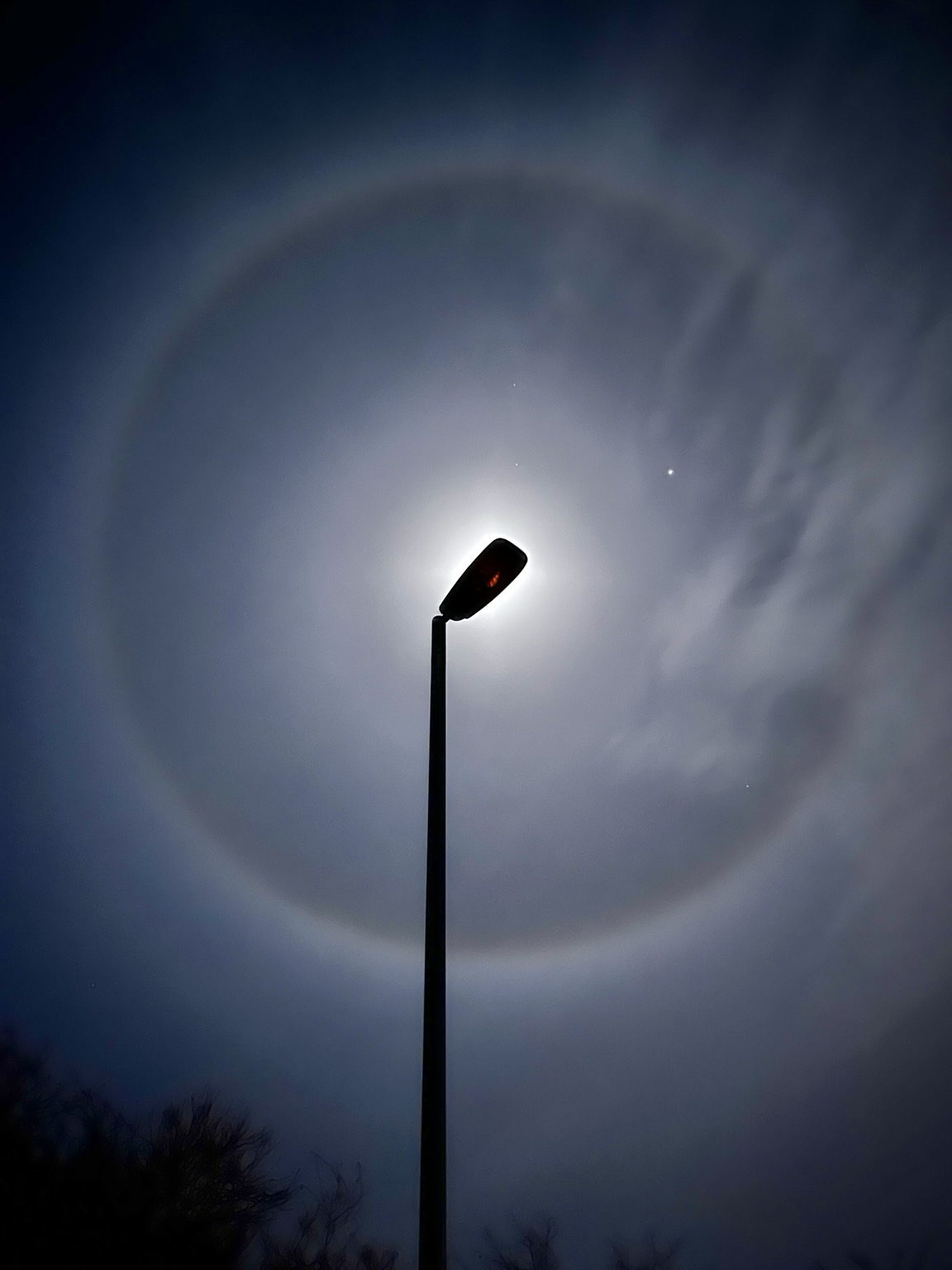 Night scene - streetlight is in the centre (it is not on/illuminated), with the lamp covering the moon, in the sky a large circle ‘halo’ circles the image, almost to the edges, it has subtle spectra, primarily just appearing as a bright white circle. Some clouds intrude into the halo in the top right, and Jupiter appears as a bright white star-like point of light at the 2 o’clock position.
