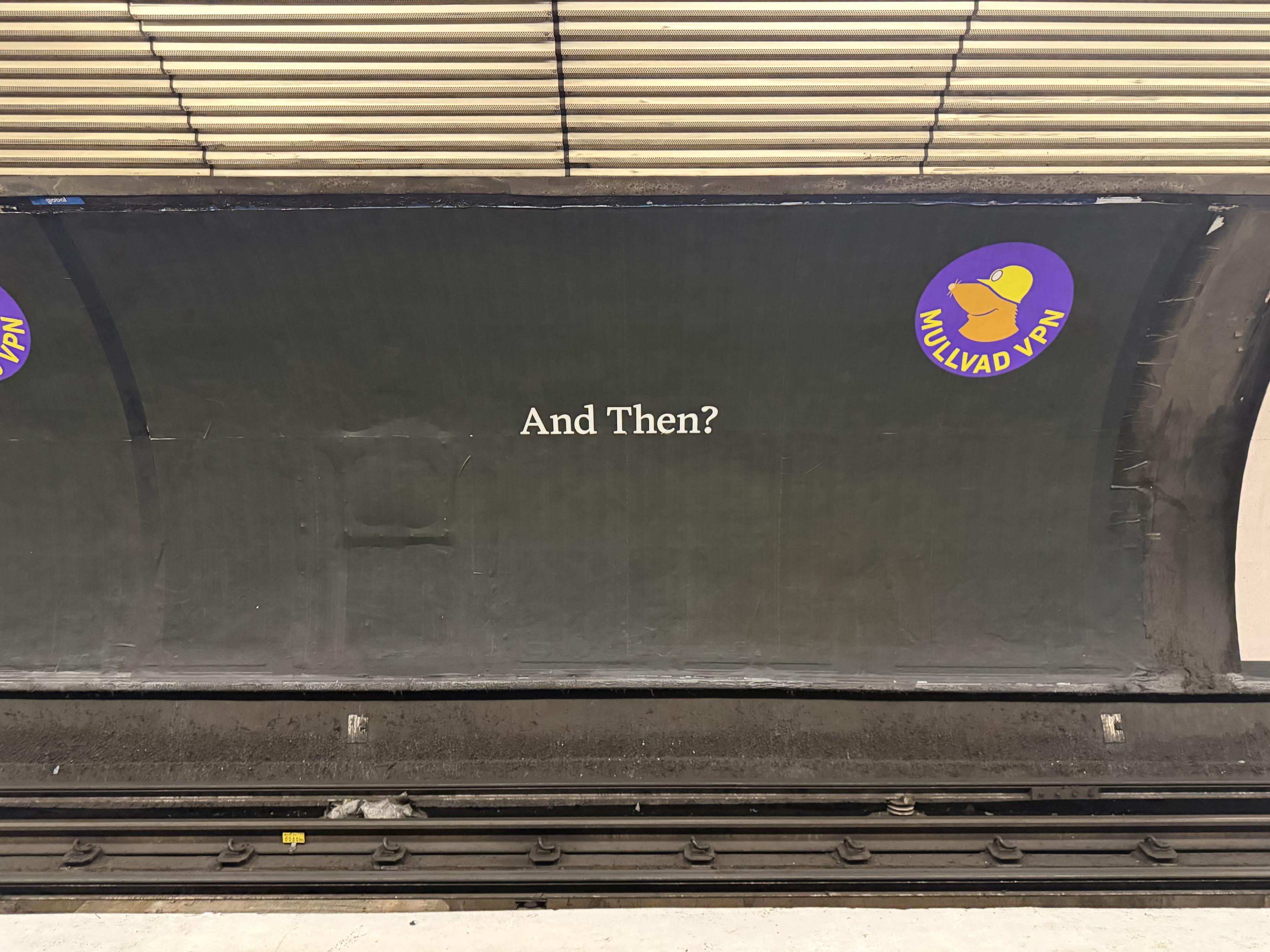 An image taken towards a Mullvad VPN advert on the side of a London underground tube station away from the platform. The image reads "And Then?" in white, across an entirely black background. The Mullvad VPN logo is in the top right corner.