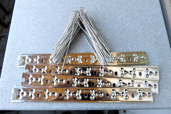 Mehrere glänzende Metallplatten liegen waagerecht übereinander auf einer hellen, leicht strukturierten Oberfläche. Die Platten sind länglich, rechteckig und spiegelnd poliert. In regelmäßigen Abständen sind symmetrische ovale Aussparungen in die Platten eingelassen. Darüber liegt ein Bündel dünner, silberfarbener Metallstäbe, die fächerförmig angeordnet sind und nach unten auf die Platten zeigen.