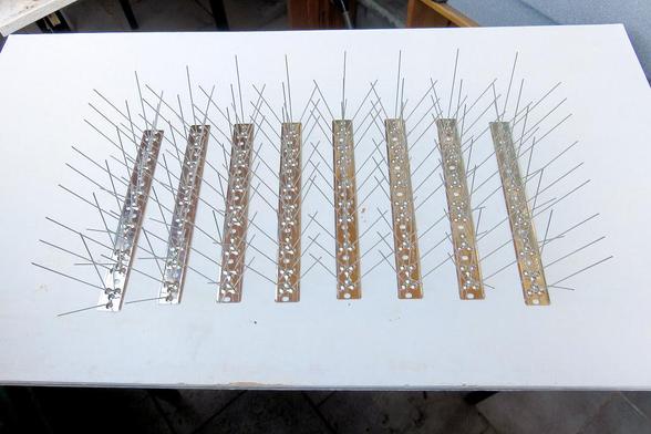 Mehrere schmale, längliche Metallleisten liegen parallel nebeneinander auf einer weißen Tischplatte. Auf jeder Leiste sind in regelmäßigen Abständen dünne, lange Metallstäbe befestigt, die schräg nach oben und zu den Seiten ragen. Die Stäbe kreuzen sich teilweise und bilden ein dichtes, spitzes Geflecht.