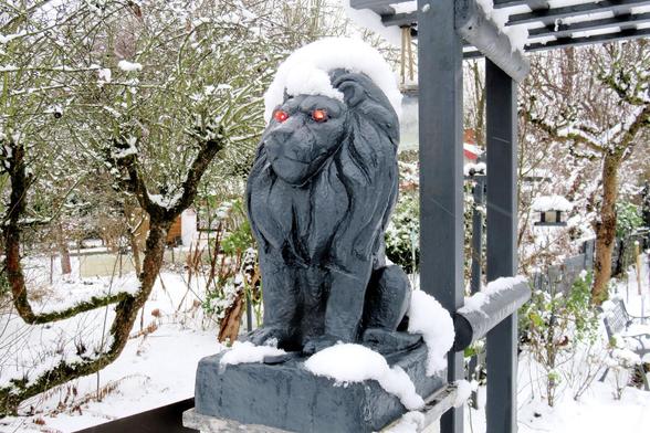 Dunkle Löwenstatue aus Stein oder Metall sitzt auf einem rechteckigen Sockel und blickt nach vorne. Auf Kopf, Rücken und Sockel liegt eine weiche Schneeschicht. Die Augen der Figur leuchten auffällig rot und heben sich deutlich vom dunklen Material ab. Es sind eingebaute LEDs. Die Mähne ist detailliert ausgearbeitet und fällt in dicken Strähnen entlang des Halses. Rechts daneben steht eine Holzkonstruktion mit Querbalken, ebenfalls mit Schnee bedeckt. Im Hintergrund sind verschneite Sträucher, …