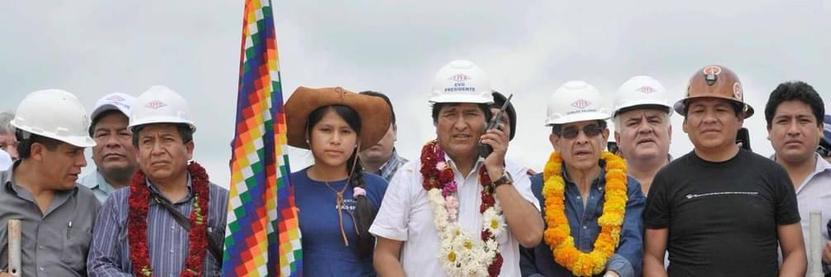 Ten Bolivian men, Evo Morales and members of his government, and one teenage girl, Cindy Sarai Vargas, pose for s propaganda photo. Most of the men are wearing hard hats. Sarai Vargas wears a cowboy hat and holds a wiphala flag. 