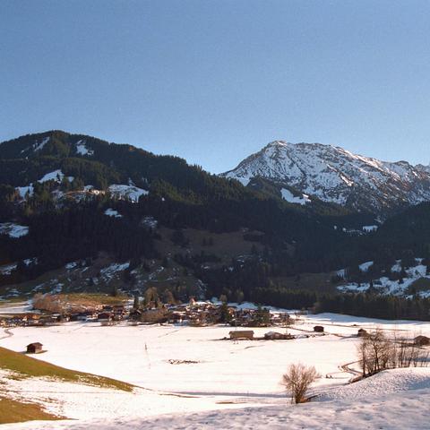 Das Bild zeigt ein breites Tal in den Alpen während des Winters. Im Vordergrund ist eine verschneite Fläche, durchzogen von einem gewundenen Weg. Einzelne Bäume und kleine Strukturen, die zu einer landwirtschaftlichen Nutzung gehören, sind über das Feld verteilt. In der Mitte des Bildes befindet sich ein kleines Dorf mit traditionellen alpinen Häusern. Die Dächer der Häuser sind mit Schnee bedeckt, was auf kürzlichen Schneefall hinweist. Im Hintergrund erheben sich bewaldete Hänge, die allmähli…