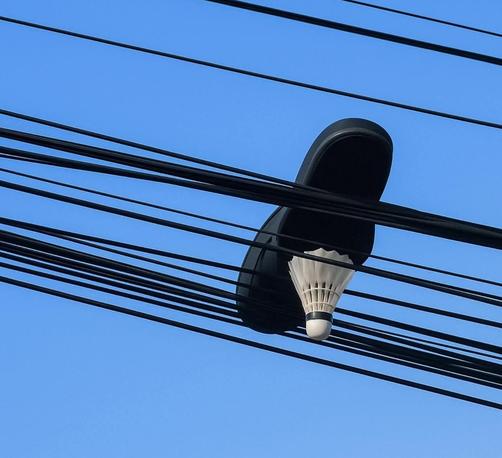 A black slide sandal and a white badminton shuttlecock trapped together in a tangle of black overhead utility wires against a clear blue sky.