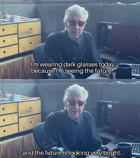 A two-panel vertical collage featuring filmmaker David Lynch in a workshop setting.

Top Panel: Lynch is seated at a desk, wearing a black button-up shirt and dark, round sunglasses. He has his signature voluminous, swept-back white hair. He is looking toward the camera with a neutral expression. White subtitle text at the bottom reads: "I'm wearing dark glasses today, because I'm seeing the future..."

Bottom Panel: A nearly identical shot of Lynch, though his expression has shifted into a…