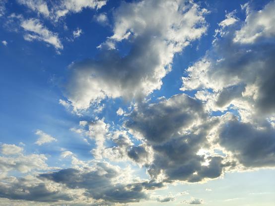 A vibrant, detailed daytime photograph of a medium-blue sky filled with a dramatic assortment of multi-layered cloud formations. Large cumulus clouds, varying in tone from bright white to darker shaded grey, fill the frame, with smaller, wispy cirrus clouds mixed throughout. Sunlight is partially obscured by a larger cloud bank on the lower-right, creating brilliant white edges on some clouds and powerful, fanned-out crepuscular sun rays (sunbeams) filtering downwards through gaps in the cloud …