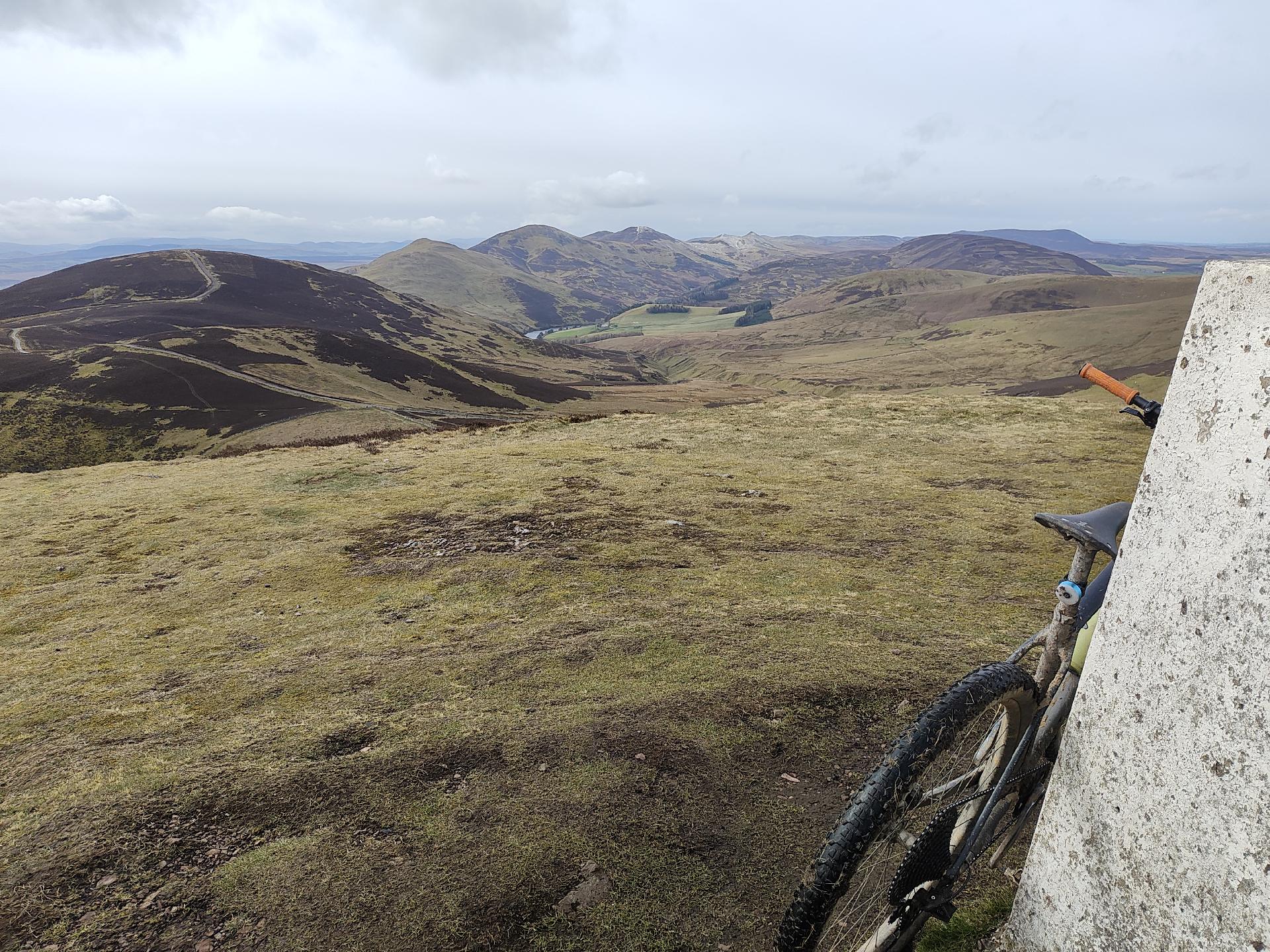 View along a range of green hills. White concrete trigpoint in foreground.