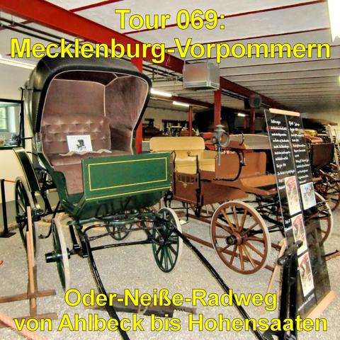 Mehrere historische Kutschen und frühe Fahrzeuge stehen in einer Ausstellungshalle. Im Vordergrund befindet sich eine grün lackierte Kutsche mit großen Speichenrädern und einem gepolsterten Innenraum. Daneben steht ein braunes, offenes Fahrzeug mit Holzrädern und Sitzbank. Weitere Kutschen sind im Hintergrund aufgereiht. Die Halle besitzt eine niedrige Decke mit sichtbaren Balken und roten Stützpfeilern. Informationstafeln stehen neben den Fahrzeugen. Der Boden ist glatt und hell.