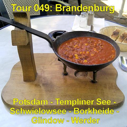 Eine kleine gusseiserne Pfanne mit langem Griff steht auf einem Holzbrett mit Halterung. In der Pfanne befindet sich ein dickflüssiger Eintopf in kräftigem Rot mit sichtbaren Bohnen und Maiskörnern. Die Pfanne steht auf drei kurzen Füßen. Daneben liegt ein Metalllöffel auf dem runden Tisch. Im Hintergrund steht ein Korb mit mehreren Stücken Brot sowie Salz- und Pfefferstreuer. Die Szene wirkt wie eine Mahlzeit im Außenbereich eines Cafés oder Biergartens.