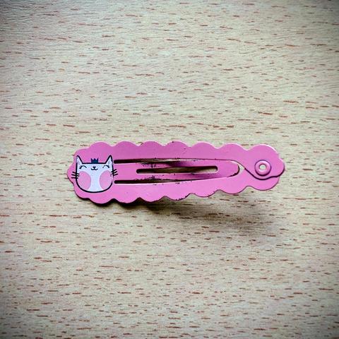 A close-up, top-down photograph of a single, worn pink metal hair snap clip resting on a light-colored wooden surface. The clip has a decorative, wavy scalloped edge. On the left end, there is a small, slightly off-center illustration of a smiling white cartoon cat face. The paint on the clip and the cat graphic is chipped and distressed, and the metal is visibly worn along the edges. The clip is oriented horizontally, with the closed snap mechanism and a tiny hole visible on the right end. The…