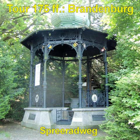 Offener Pavillon aus dunklem, kunstvoll verziertem Gusseisen inmitten dichter grüner Vegetation. Die achteckige Konstruktion steht auf einem steinernen Sockel mit eingelassenen Tafeln. Schlanke Säulen tragen ein reich geschmücktes Dach mit filigranen Ornamenten und einem kuppelartigen Abschluss. Zwischen den Säulen befinden sich dekorative Gitterelemente und kleine Wappen. Umgeben ist der Pavillon von hohen Bäumen und Sträuchern, die das Bauwerk teilweise einrahmen. Der Boden davor ist gepflast…