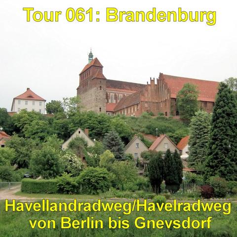 Auf einem bewaldeten Hügel erhebt sich ein großes historisches Backsteingebäude mit roten Ziegeldächern. Ein massiver Turm mit kleinem Aufsatz ragt über den Komplex hinaus. Das langgestreckte Hauptgebäude besitzt hohe, schmale Fenster und ein steiles Dach. Unterhalb des Hügels stehen mehrere Wohnhäuser mit hellen Fassaden und roten Dächern, umgeben von dichten Gärten und Bäumen. Hohe Nadel- und Laubbäume rahmen die Szene ein. Im Vordergrund verläuft eine Wiese mit einem schmalen Weg. Der Himmel…