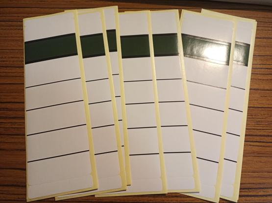 Mehrere weiße Etiketten mit dunkelgrünem Balken liegen fächerförmig auf einer Holzoberfläche.