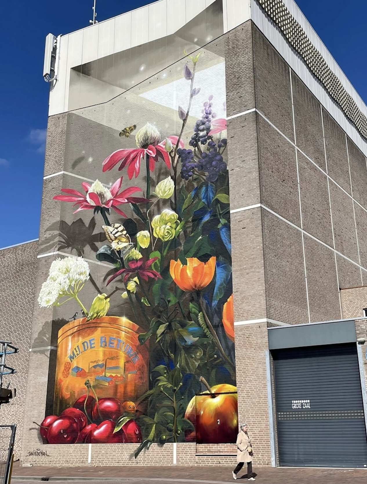 Towering 3D illusion floral mural by Jan Is De Man in Tiel, Netherlands. Oversized wildflowers, cherries, apples, and a vintage Betuwe fruit crate are beautifully painted on a tall theater wall.