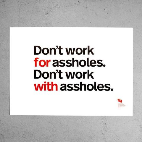 A white poster with four lines of centered text hangs on a textured gray wall. The top line reads, “Don’t work” in black letters. The second line says, “for assholes.”, with “for” in red and “assholes.” in black. The third line reads, “Don’t work” in black letters. The fourth line says, “with assholes.”, with “with” in red and “assholes.” in black. In the lower right corner, there is a small red logo of stacked shipping boxes and very small text.