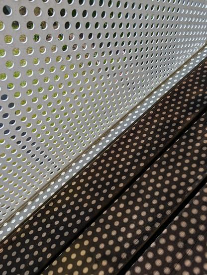 A detailed, angled close-up photo shows a white metal balcony panel perforated with dense rows of circular holes, like a polka-dotted structure. The panel slopes from the top-left towards the bottom-right, where it meets dark wooden deck planks at an angle. Strong sunlight from the upper-right shines through the panel's holes, casting a pattern of small, bright white dots on the dark wood decking, matching the perforated design. Through the holes in the upper panel, a blurry view of a green gra…