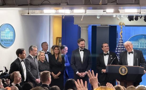 A group of people in formal attire standing behind a podium with the presidential seal in a White House press briefing room, with an audience clapping in the foreground and a blue curtain, American flag, and White House emblem in the background.