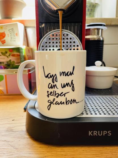 Eine rote Krups-Kapselmaschine brüht gerade einen Strahl frischen Kaffee in eine weiße Tasse, die auf dem silbernen Abtropfgitter steht. Auf der Tasse ist in schwarzer, geschwungener Handschrift der Spruch „lass mal an uns selber glauben...“ aufgedruckt. Das Gerät steht auf einer hölzernen Arbeitsplatte. Im leicht unscharfen Hintergrund sind links gestapelte Teeschachteln und rechts eine kleine weiße Dose mit Deckel zu sehen, beleuchtet von warmem Sonnenlicht.