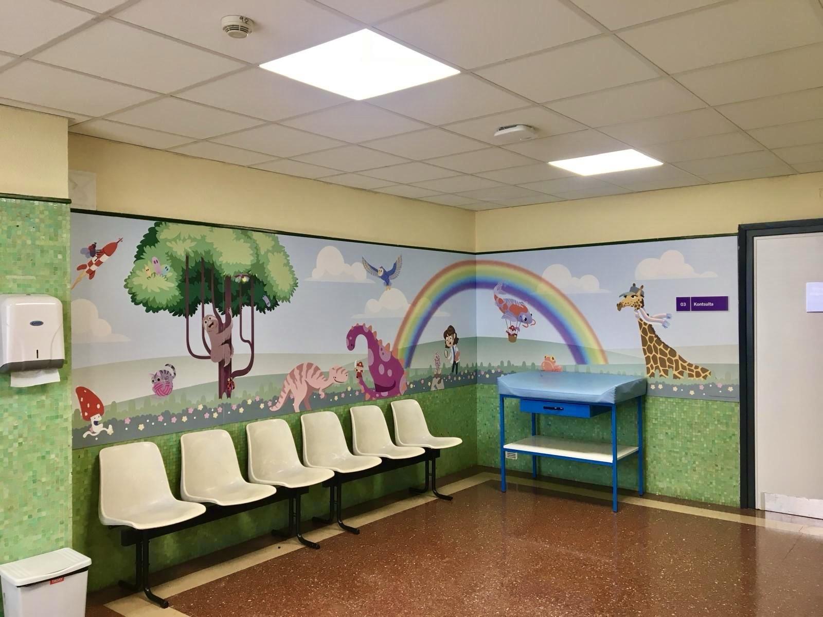 The image shows a brightly colored children's waiting area featuring a mural with playful animal illustrations, a rainbow, and nature scenes. There are several white chairs for seating and a blue cart against the wall. The overall atmosphere appears cheerful and inviting.