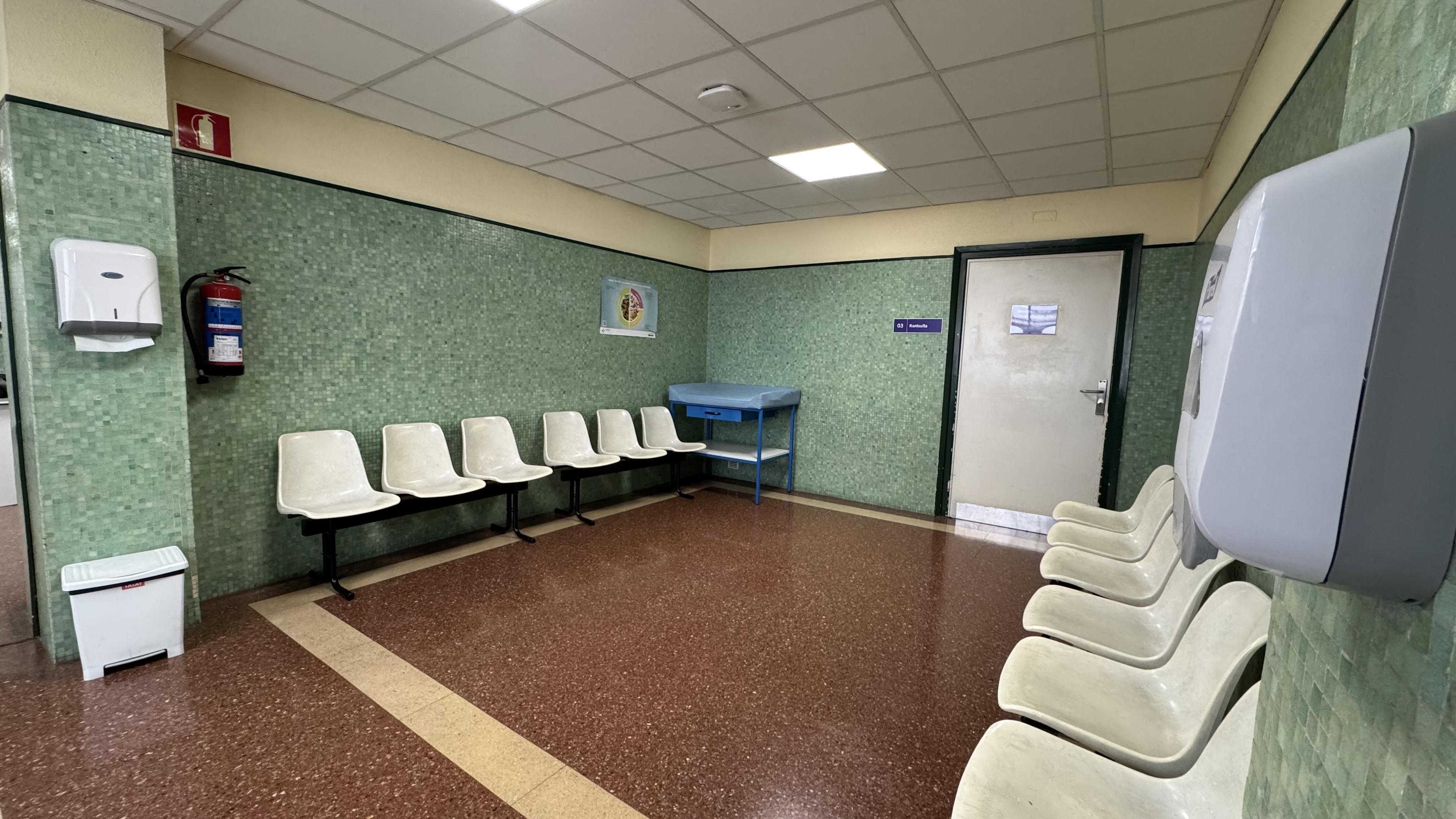 A waiting area in a medical facility featuring green tiled walls, several white plastic chairs arranged against the wall, a blue examination table, a hand sanitizer dispenser, and a fire extinguisher. The floor is a mix of brown and beige tiles.