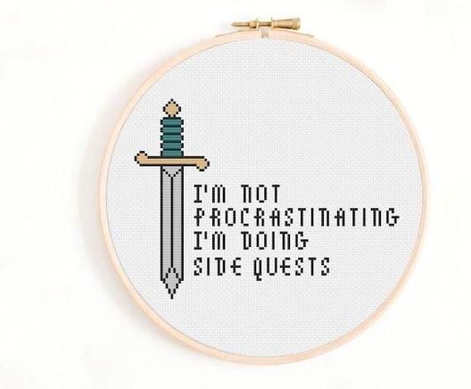 A circular wooden embroidery hoop on a plain white background displays a cross-stitch design on white Aida fabric. The left side features a cross-stitch sword with a teal-striped hilt, gold crossguard and pommel, and a grey and silver blade. To the right of the sword, the cross-stitch text reads: “I’M NOT PROCRASTINATING I’M DOING SIDE QUESTS” in black, pixelated capital letters.
