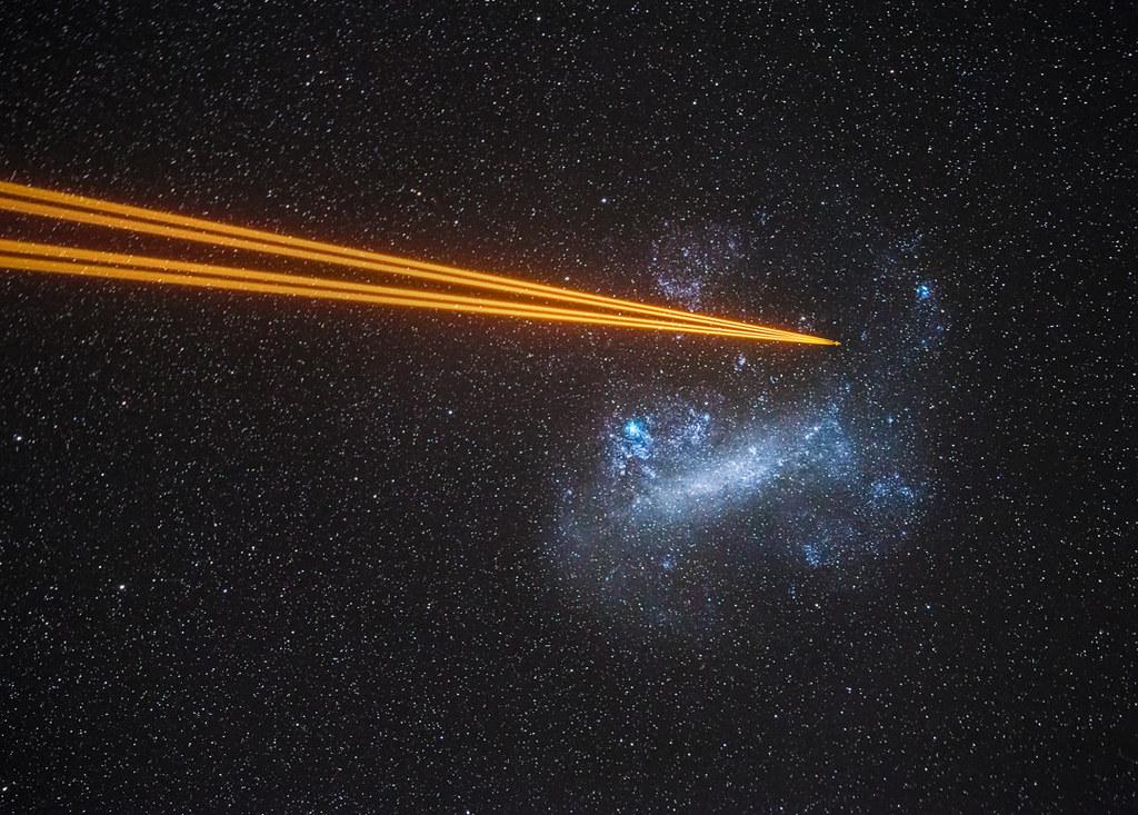 A long exposure image of the night sky, showing a blue cloud-like galaxy sparkled with stars. Four orange laser beams come from the left, seemingly converging towards a location within the galaxy.