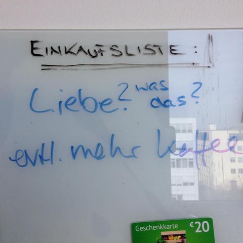 Eine Nahaufnahme einer weißen Magnetwand oder Glasoberfläche, auf der handschriftliche Notizen mit Markern stehen. Oben steht in Großbuchstaben mit schwarzem Marker das Wort „EINKAUFSLISTE:“, das doppelt unterstrichen ist.

Darunter befinden sich zwei Einträge in blauer Handschrift:

Zuerst das Wort „Liebe?“, daneben steht etwas kleiner „Was ist das?“.

Darunter folgt der Satz „evtl. mehr Kaffee“.

Am unteren rechten Rand ist die Ecke einer grünen Geschenkkarte im Wert von 20 € zu sehen…