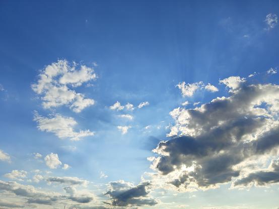 A wide-angle, low-angle shot of a vibrant blue sky filled with scattered white and grey cumulus clouds. In the lower right, a large, dark cloud partially obscures the sun, causing bright crepuscular rays (sunbeams) to radiate upward and outward into the clear blue expanse. Small, wispy cloud fragments are scattered across the center, and a faint, thin vapor trail from a plane is visible in the middle left. The overall lighting is bright and ethereal, suggesting late afternoon.